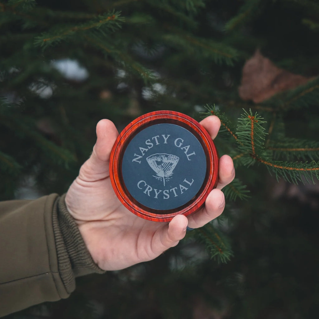 Hand holding a container labeled 'Nasty Gal Crystal' against a blurred natural background