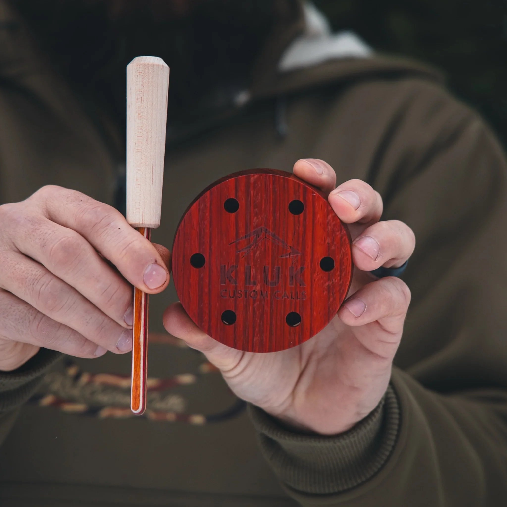 Person holding a wooden tool and a red circular object with 'Kluk' branding.