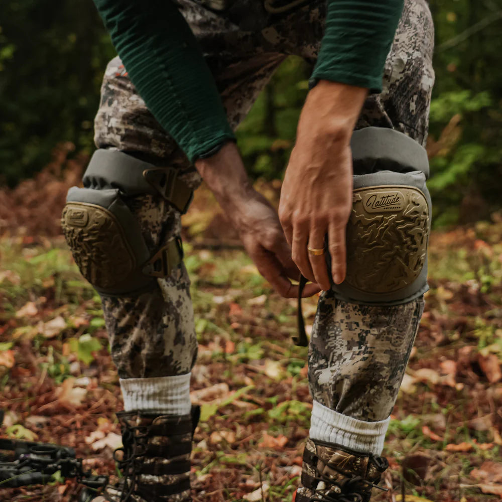 Person wearing camouflage pants and knee pads in a forest setting