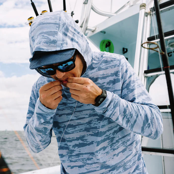 guy wearing a blue camouflage hoodie on a boat while biting fishing line  