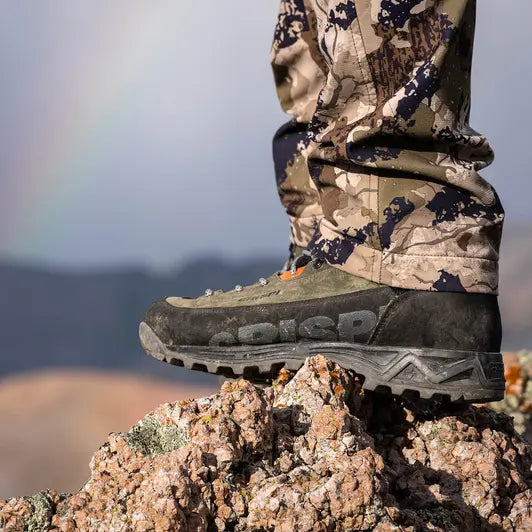 Person wearing camouflage pants and hiking boots on a rocky mountain with a rainbow in the background