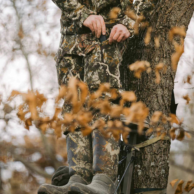 Person in camouflage clothing and boots standing near a tree in a forest setting.