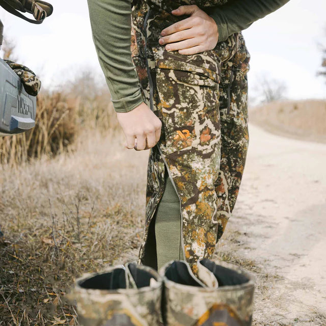 Person in camouflage hunting attire with boots and a rifle case on a dirt path.