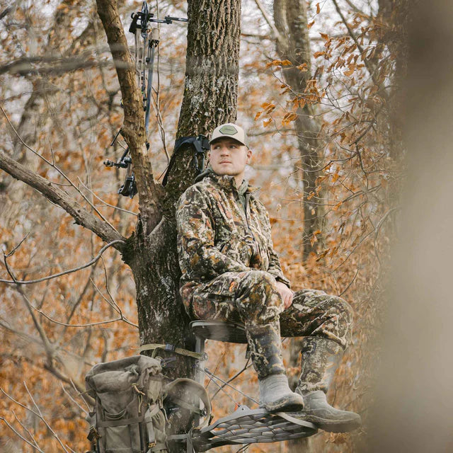 Man in camouflage sitting in a tree stand in a forest during autumn.