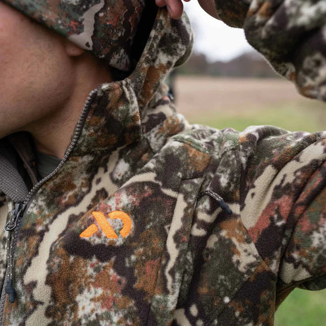 Person wearing a camouflage jacket with a brand logo, sitting outdoors.