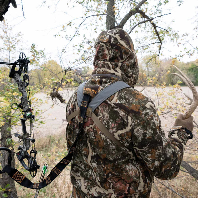 Hunter in camouflage gear with a bow, holding a deer in a forest setting