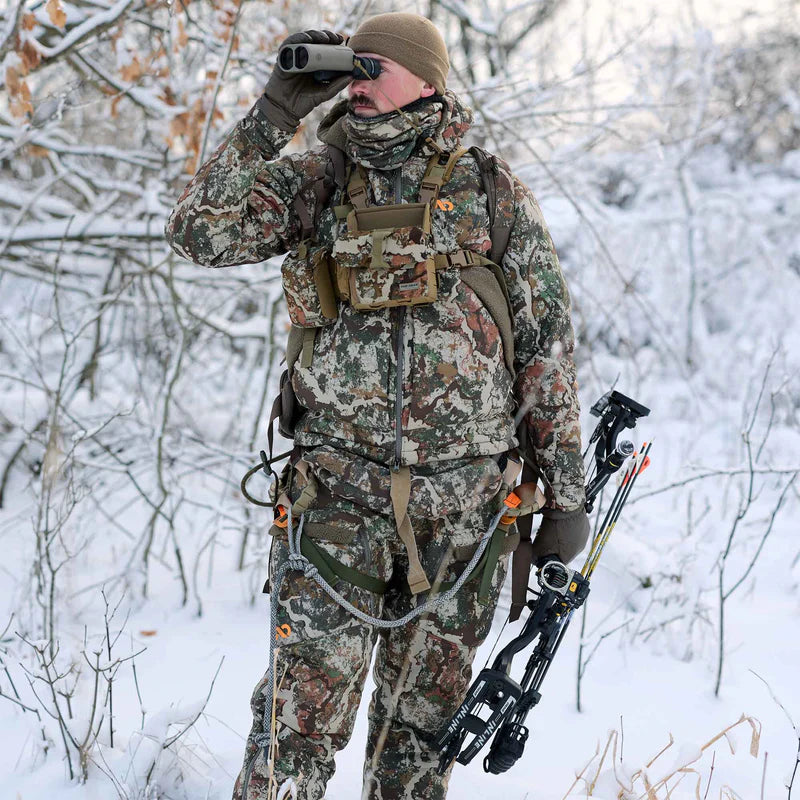Person in camouflage hunting gear with binoculars in a snowy forest