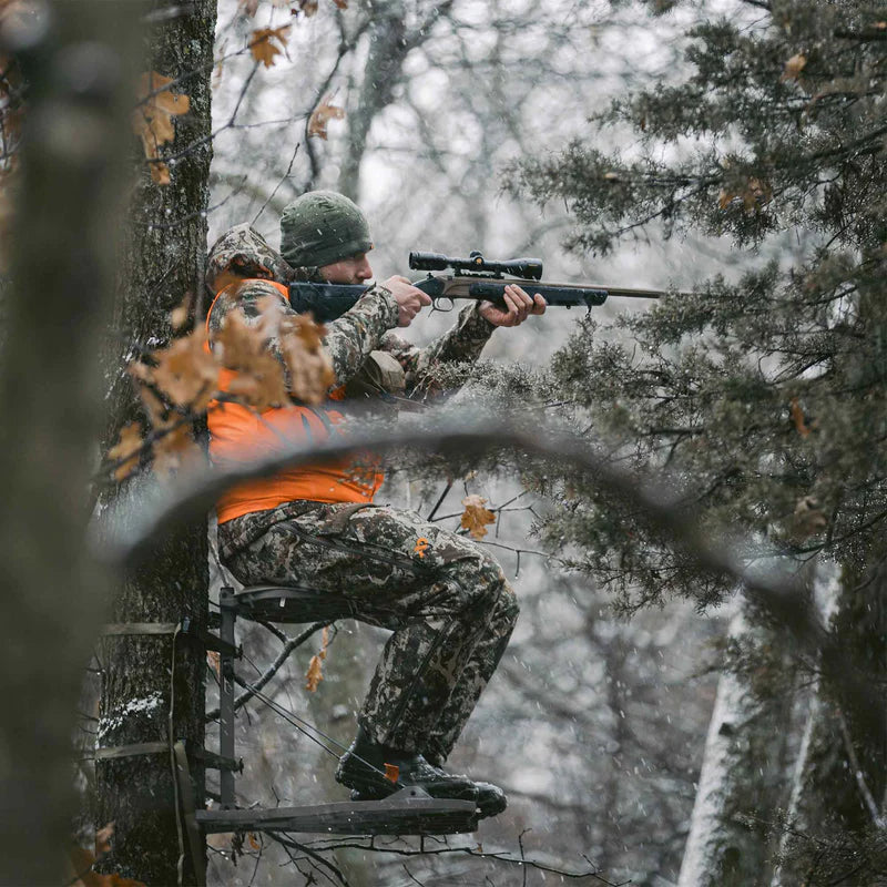 Hunter in camouflage and orange vest using a rifle in a snowy forest.