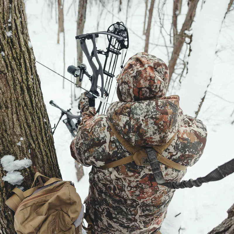 Hunter in camouflage gear with a bow, standing in a snowy forest.