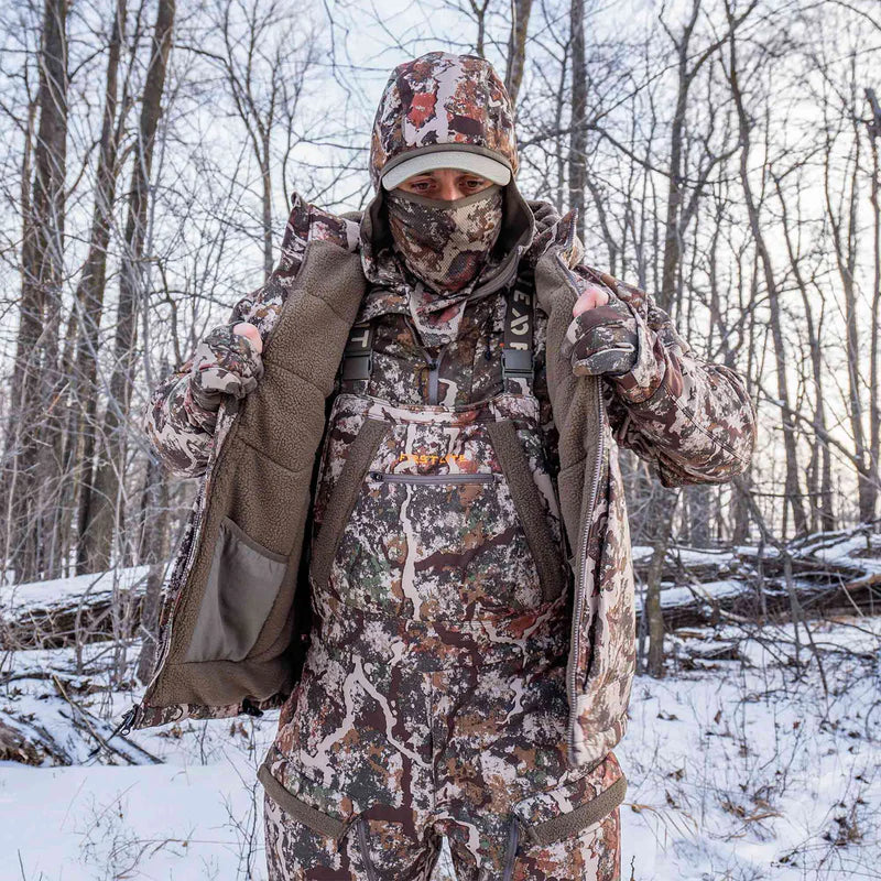 Person in full camouflage hunting gear standing in a snowy forest.