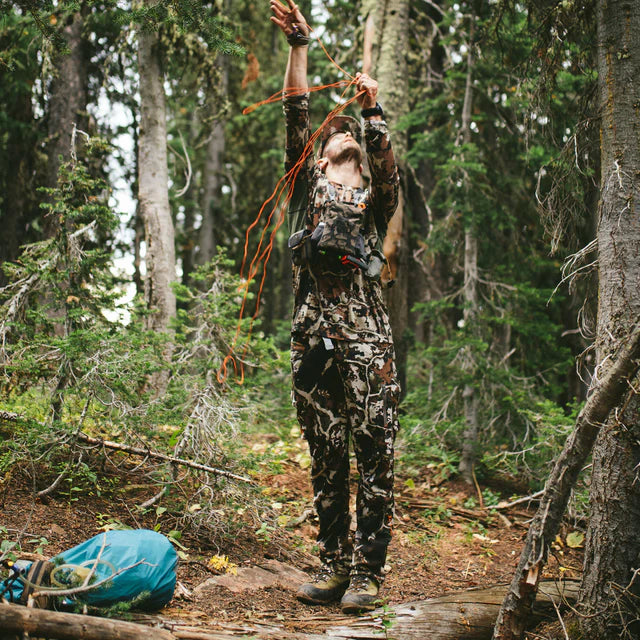 Person in camouflage gear in a forest setting with a bow and arrow.