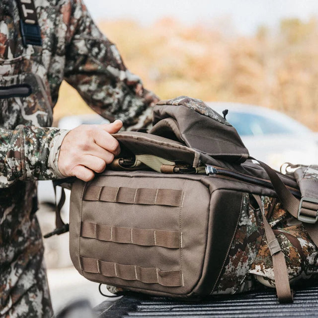Person in camouflage holding a brown tactical bag outdoors