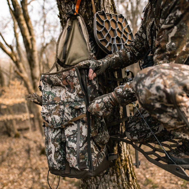Hunter in camouflage gear using a tree stand with a backpack and archery equipment.
